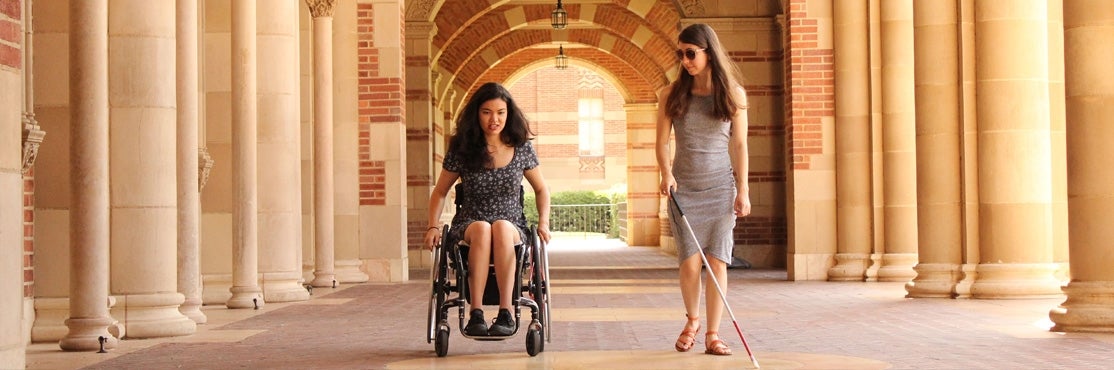 A person in a wheelchair and a person holding a white cane proceed side by side on the UCLA campus 