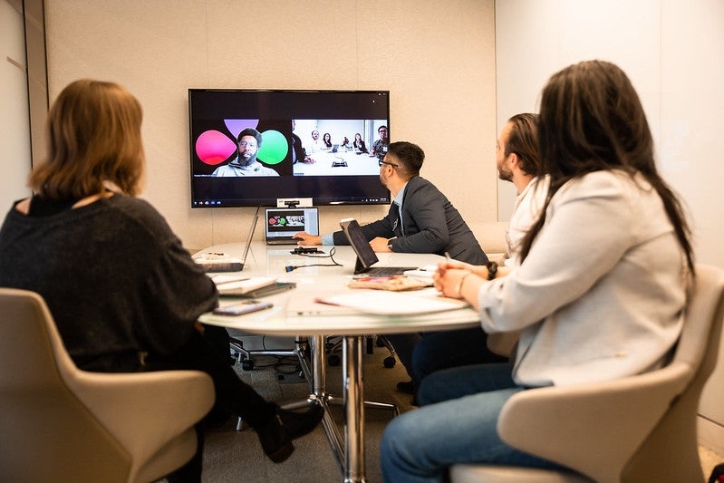 A group of MBA students seated around a conference table with additional figures on zoom on a wallmounted monitor